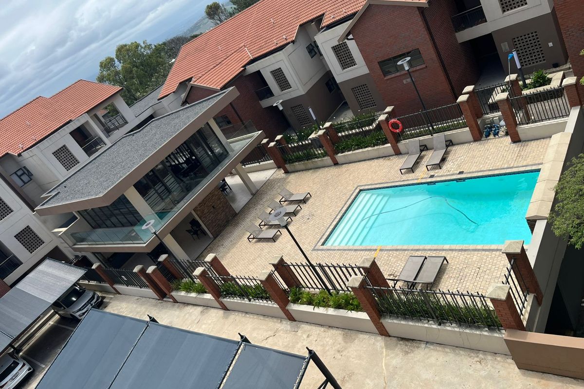 An elevated view of a communal apartment complex pool area featuring a paved deck, brick boundary walls, and surrounding residential buildings.