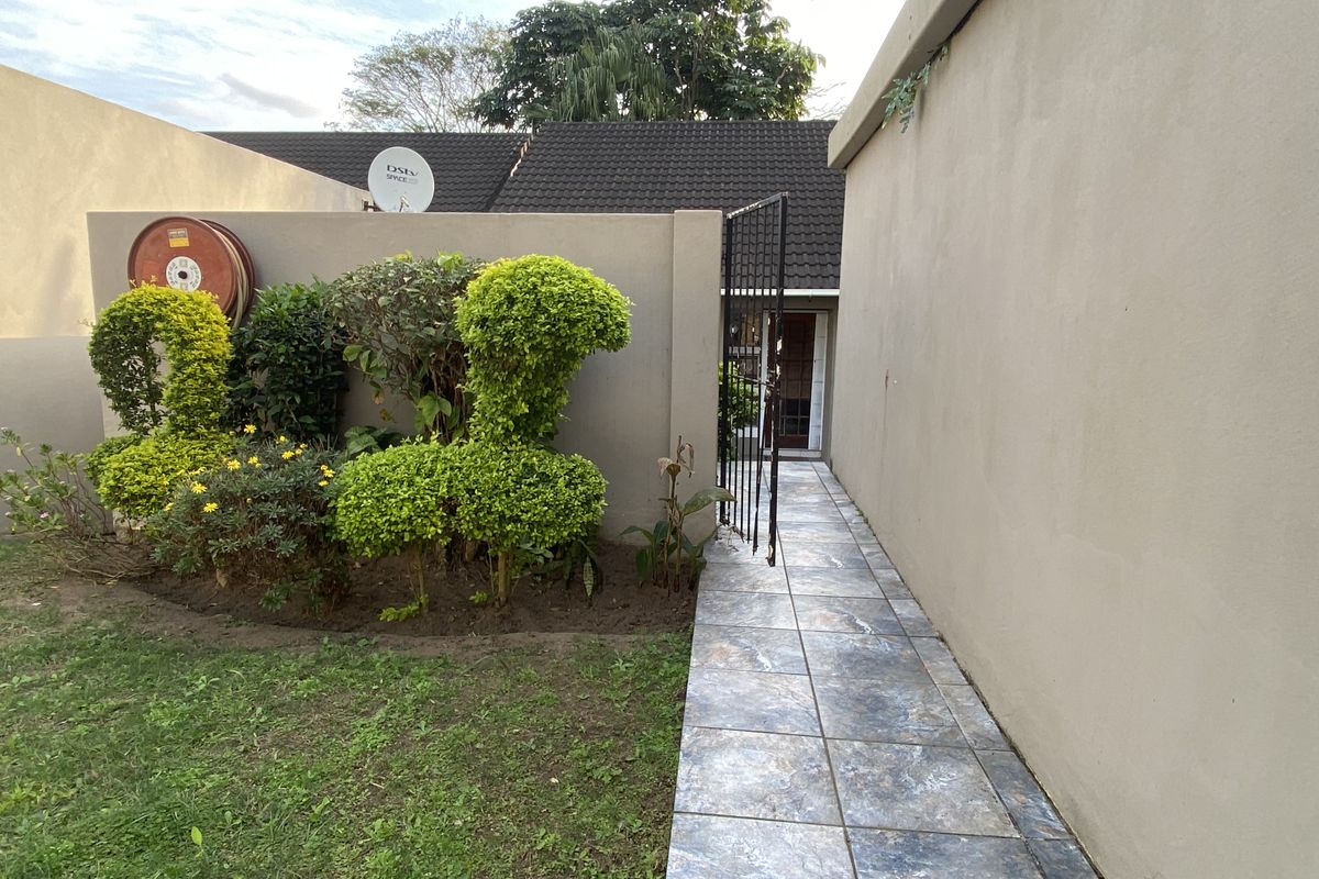 A rear exterior view of the townhouse featuring a tiled walkway leading to a gated entrance, bordered by a boundary wall and garden area.