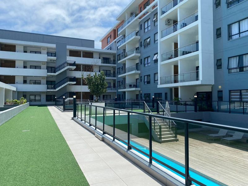 Modern apartment complex featuring a communal swimming pool, glass balustrades, artificial lawn areas, and paved walkways between multi-story residential buildings.
