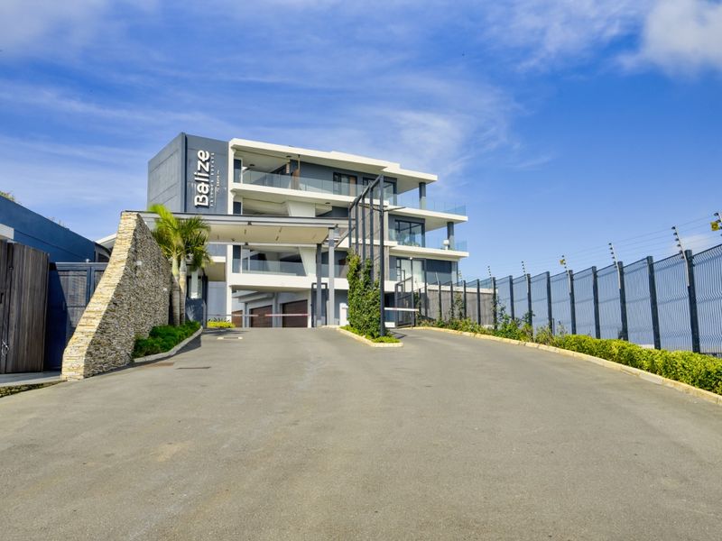 Modern apartment complex entrance featuring a paved driveway, secure gated access, and contemporary architectural design with glass-fronted balconies.