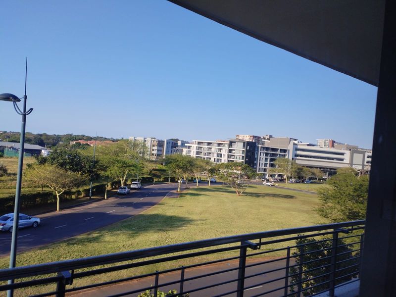 An elevated view overlooking a suburban street, green space, and surrounding apartment buildings, captured from a balcony with a metal balustrade.