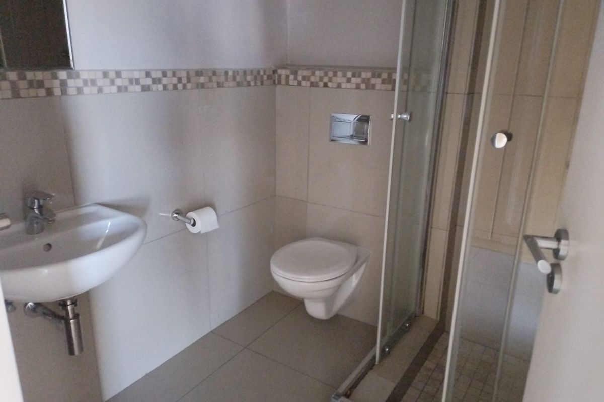 A clean, modern bathroom featuring a wall-mounted toilet, glass shower enclosure, and a pedestal basin with neutral wall tiling and a decorative mosaic border.