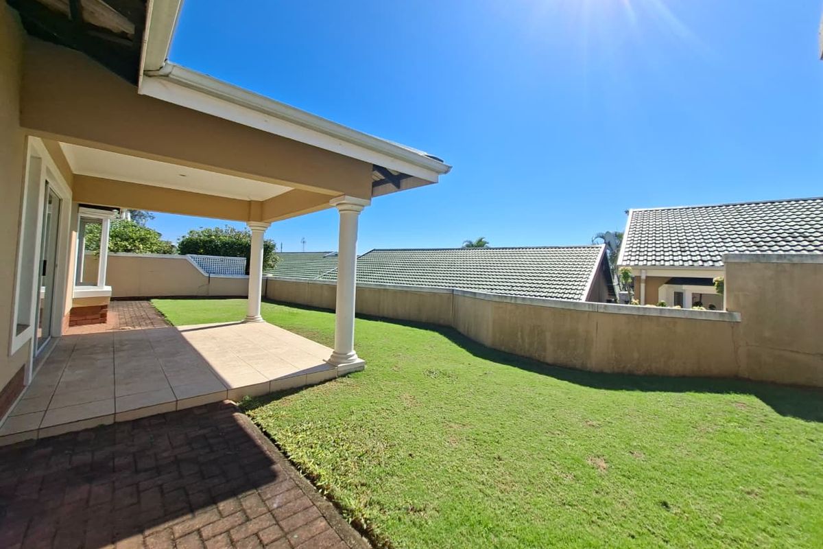 A spacious rear garden featuring a covered patio with tiled flooring, concrete columns, and a well-maintained lawn enclosed by a boundary wall.