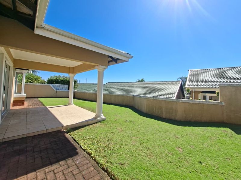 A spacious rear garden featuring a covered patio with tiled flooring, concrete columns, and a well-maintained lawn enclosed by a boundary wall.