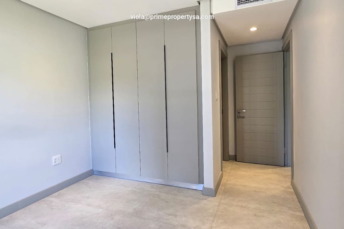 A modern, empty bedroom featuring neutral tiled flooring, built-in cupboards with sleek handles, and recessed ceiling lighting.
