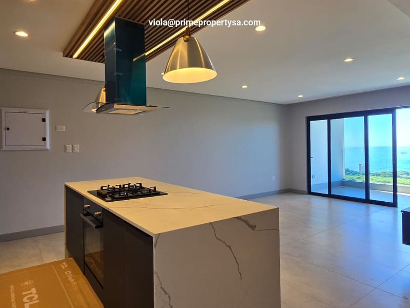 Modern open-plan kitchen and living area featuring a stone-topped island, built-in appliances, and large sliding doors leading to a balcony with ocean views.