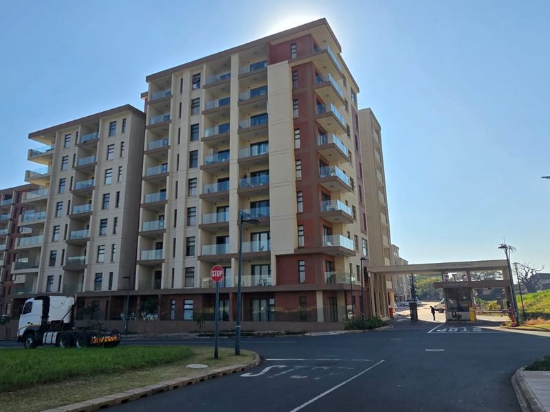 A modern multi-story apartment complex featuring glass balconies and a secure gated entrance, viewed from the street level.