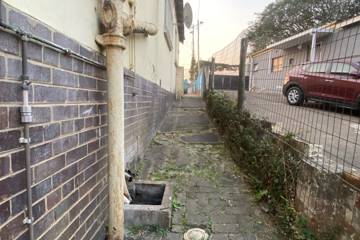 A narrow side exterior walkway featuring brick walling, a corroded drainage pipe, and uneven paved surfaces requiring maintenance.