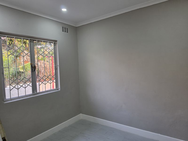 A clean, empty bedroom featuring neutral grey walls, recessed downlighting, and a window fitted with a security gate.