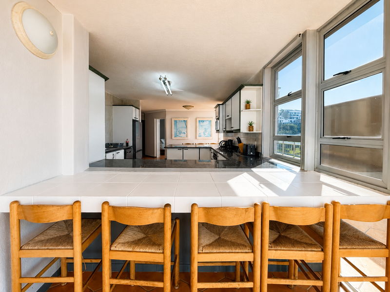 Open-plan kitchen and dining area featuring a tiled breakfast bar, granite countertops, and ample built-in cabinetry with views through large aluminium windows.