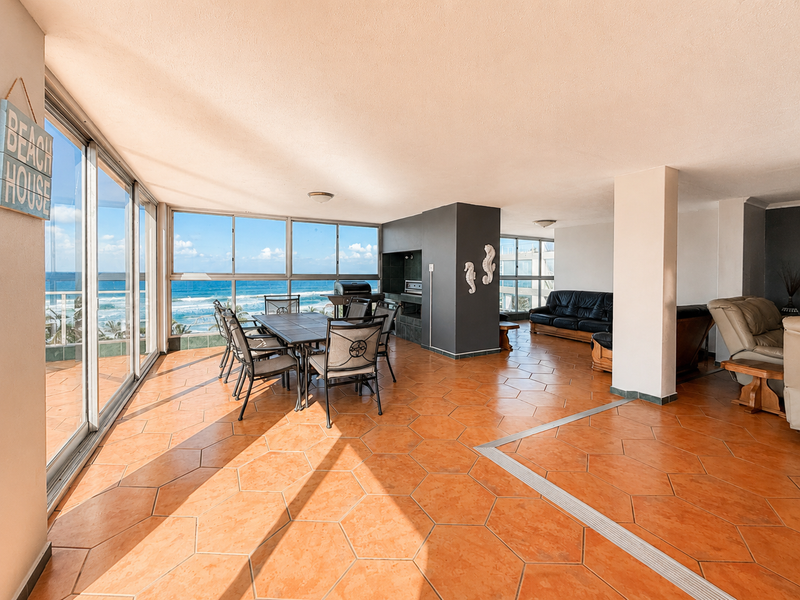 Spacious open-plan living and dining area featuring terracotta floor tiles, a built-in braai, and expansive sliding glass doors with panoramic ocean views.