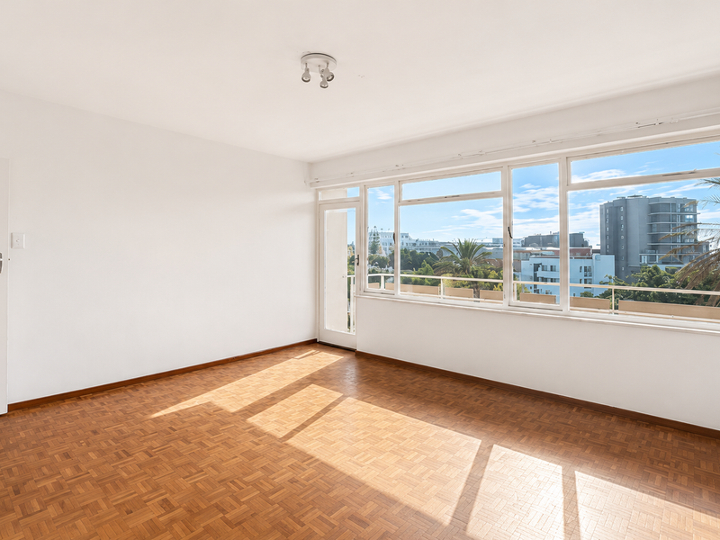 A bright, empty living room featuring classic parquet flooring, large windows with urban views, and direct access to a balcony.