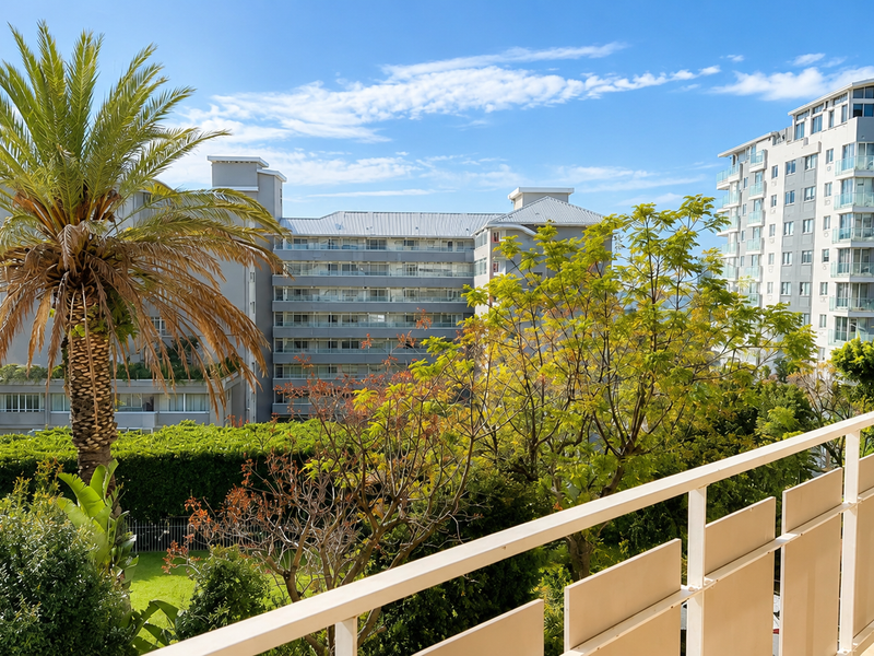 A scenic elevated view from a balcony overlooking lush greenery, a swimming pool, and surrounding apartment buildings under a clear blue sky.