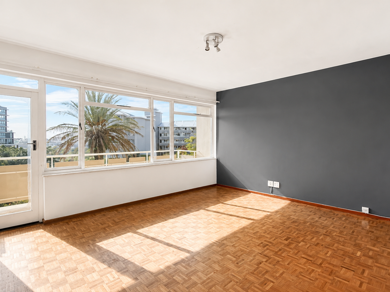 Bright and spacious living area featuring classic parquet flooring, a striking dark accent wall, and large windows providing ample natural light and balcony access.