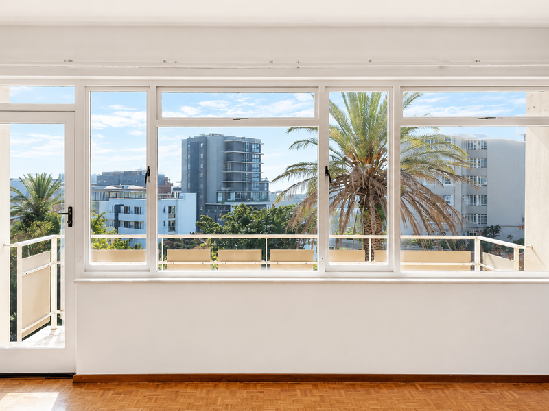 A bright, empty living room featuring classic parquet flooring, large windows, and direct access to a balcony with urban views.