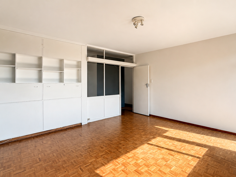 A bright, empty living room featuring classic parquet flooring, a large built-in white shelving unit, and an internal glass partition wall.