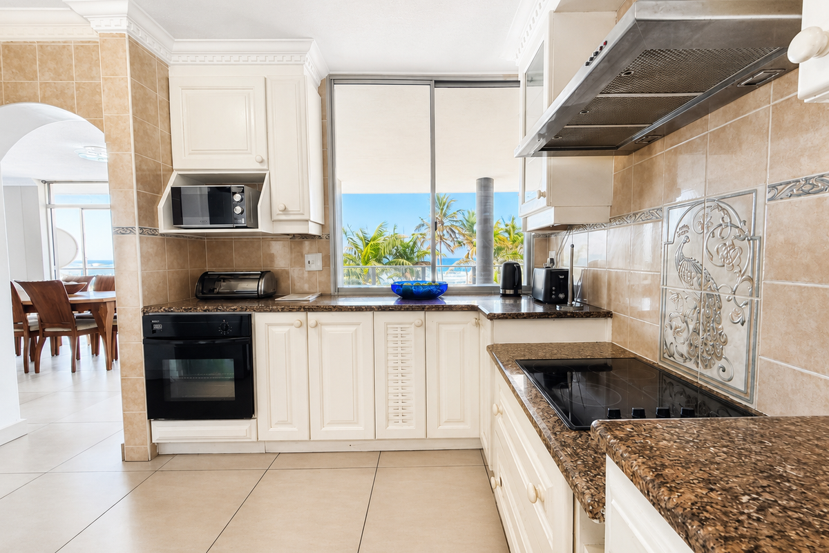 A well-maintained kitchen featuring granite countertops, built-in appliances, tiled splashbacks, and crown molding, with a view through the window to the exterior.