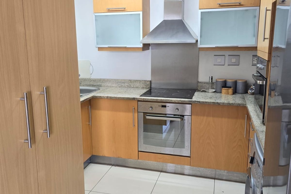 A functional kitchen featuring light wood cabinetry, granite countertops, a built-in oven, electric hob, and stainless steel extractor fan with tiled flooring.
