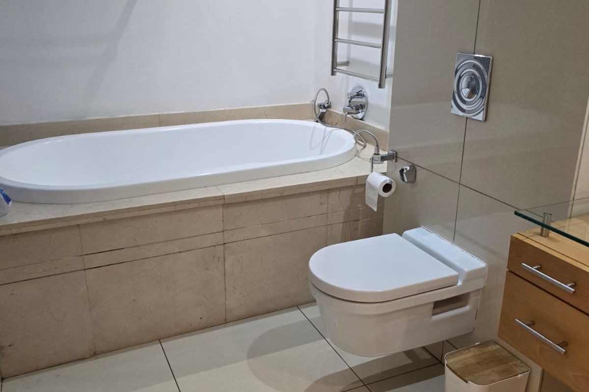 Modern bathroom featuring a built-in bathtub, wall-mounted toilet with concealed cistern, heated towel rail, and a vanity cabinet with a glass countertop.