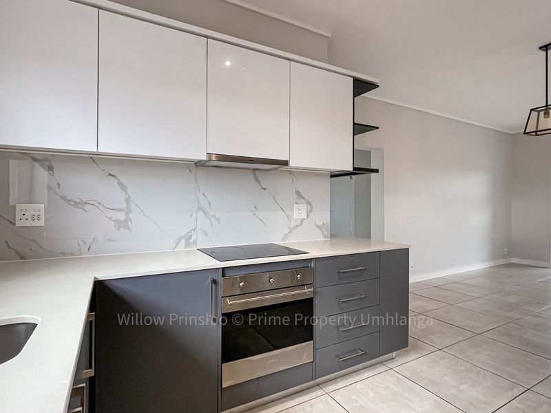 Modern open-plan kitchen featuring sleek two-tone cabinetry, stone countertops, marble-look splashback, and integrated appliances, flowing into a bright living area with tiled floors.