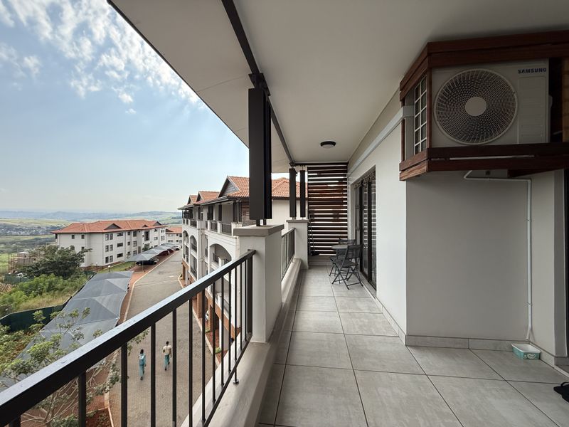 A spacious covered balcony featuring tiled flooring, a metal balustrade, and an integrated air conditioning unit, offering views over the apartment complex.