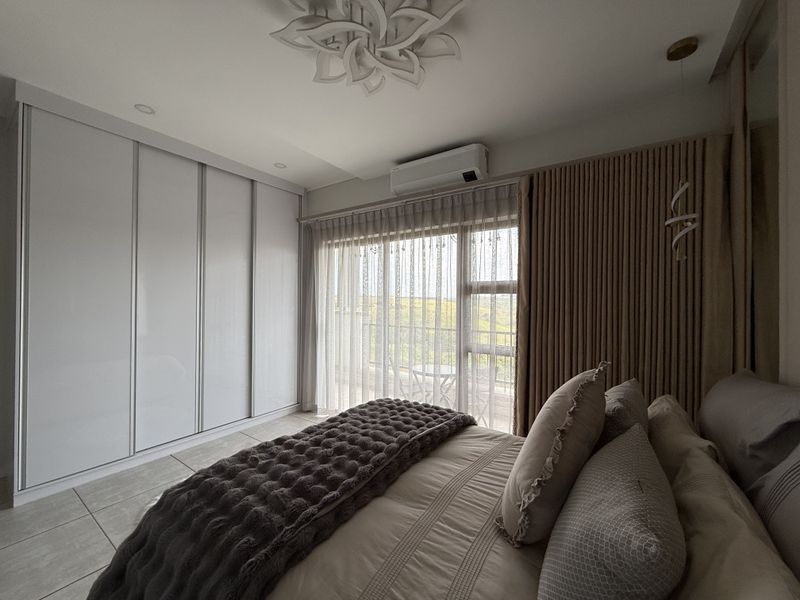 A modern, well-maintained bedroom featuring built-in sliding wardrobes, tiled flooring, a split-system air conditioner, and direct access to a private balcony.