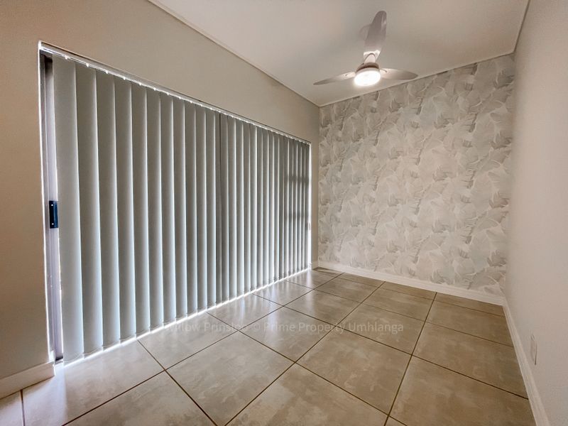 A bright, empty bedroom featuring large sliding glass doors with vertical blinds, tiled flooring, a ceiling fan, and a decorative feature wall.