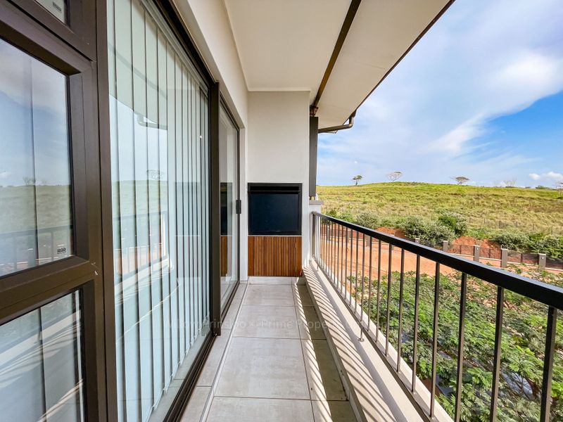 A clean, covered balcony featuring tiled flooring, a built-in braai, and a metal balustrade, offering scenic views of the surrounding landscape.