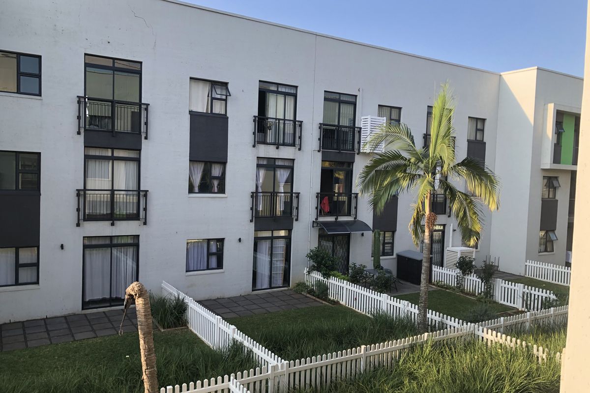 Rear view of a multi-story apartment complex featuring private ground-floor gardens with white picket fencing, paved patios, and upper-level balconies.