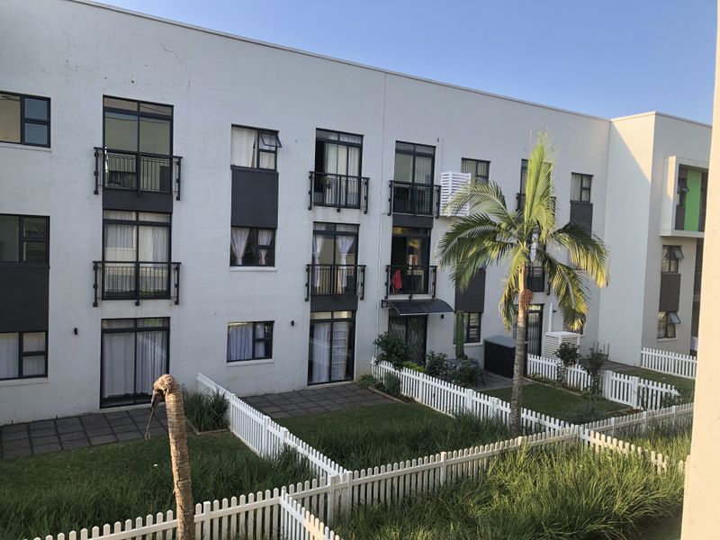 Rear view of a multi-story apartment complex featuring private ground-floor gardens with white picket fencing, paved patios, and upper-level balconies.