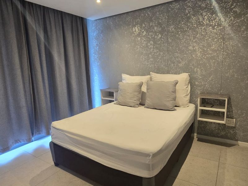 A modern bedroom featuring tiled flooring, recessed ceiling lighting, and a textured feature wall with integrated wall-mounted bedside shelving.