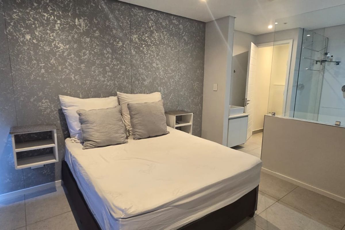 Modern bedroom featuring a textured feature wall, tiled flooring, and an open-plan en-suite bathroom with a glass-enclosed shower.