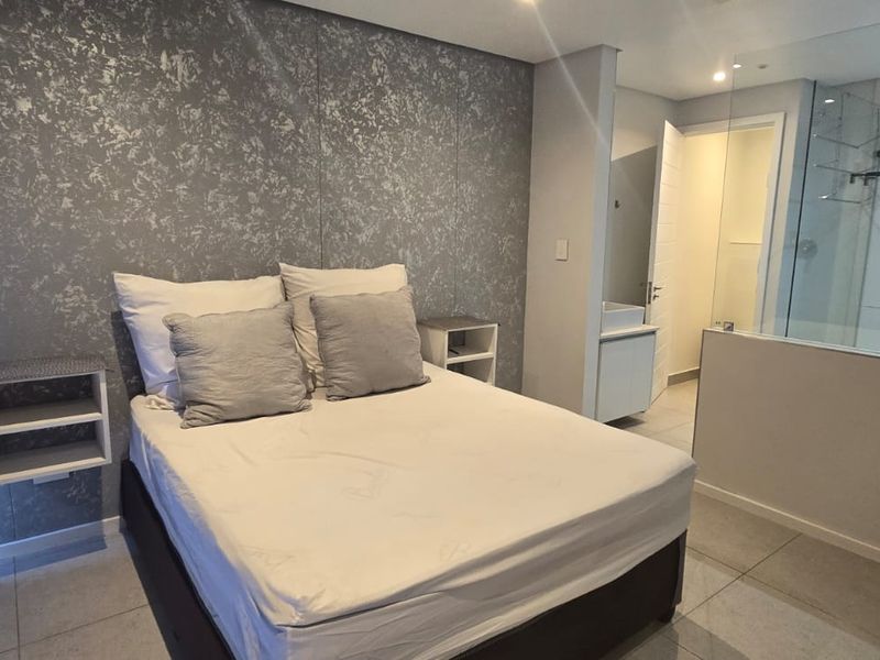 Modern bedroom featuring a textured feature wall, tiled flooring, and an open-plan en-suite bathroom with a glass-enclosed shower.
