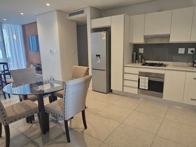 Modern open-plan living area featuring a sleek kitchen with built-in appliances, tiled flooring, and recessed lighting, seamlessly flowing into the dining and lounge space.