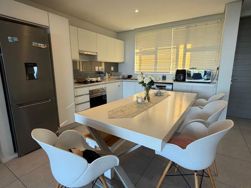 Modern open-plan kitchen and dining area featuring sleek white cabinetry, integrated appliances, tiled flooring, and ample natural light through venetian blinds.