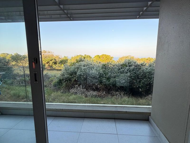 A clean, modern balcony featuring tiled flooring, a glass balustrade, and a sliding door, offering a peaceful view of the surrounding greenery.