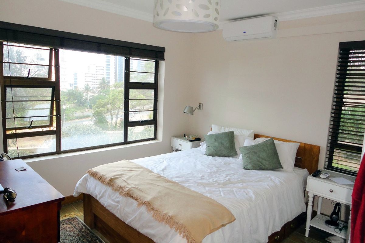 A well-lit bedroom featuring tiled flooring, a split-system air conditioner, and large windows with venetian blinds offering an urban view.