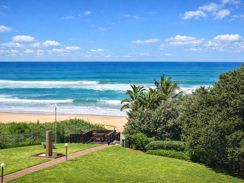 A scenic view of the ocean from a well-maintained garden featuring a paved walkway, outdoor shower, and direct beach access via a wooden deck.