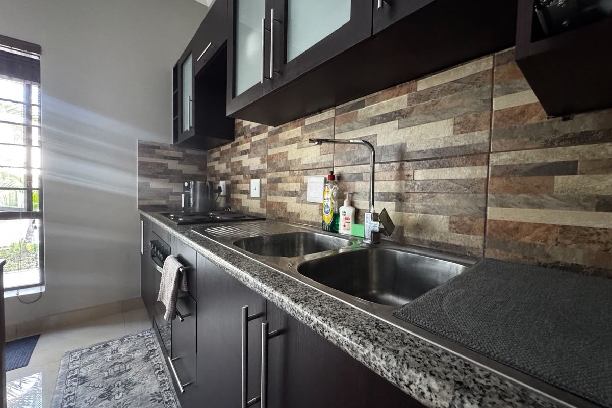 A functional kitchen featuring dark wood cabinetry, granite countertops, a double stainless steel sink, and a decorative stone tile backsplash.