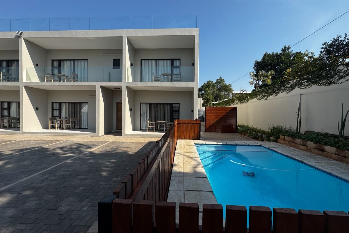 Modern apartment complex exterior featuring a swimming pool, paved parking area, and private balconies with glass balustrades.