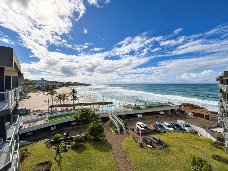 Elevated coastal view overlooking the apartment complex grounds, swimming pool, parking area, and the adjacent sandy beach and ocean.