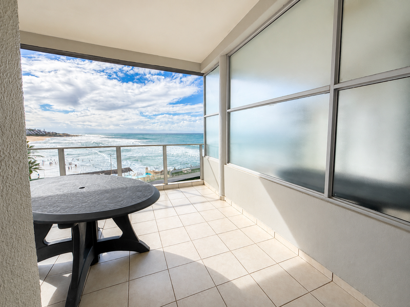 A spacious covered balcony featuring tiled flooring, a glass balustrade, and a frosted glass privacy screen, offering expansive ocean views.