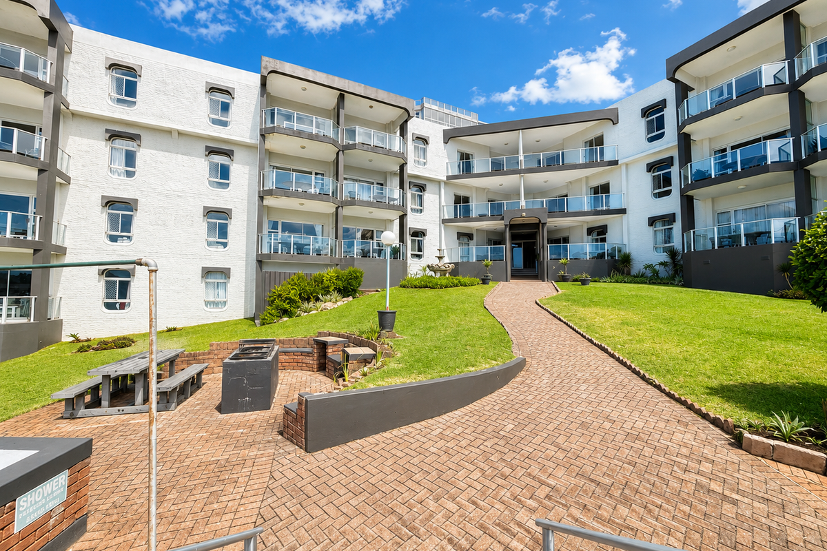Rear view of the apartment complex featuring a landscaped lawn, brick paved pathways, built-in braai area, and multiple balconies with glass balustrades.