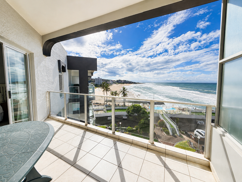 A spacious covered balcony featuring tiled flooring and a glass balustrade, offering expansive panoramic views of the coastline and beach.