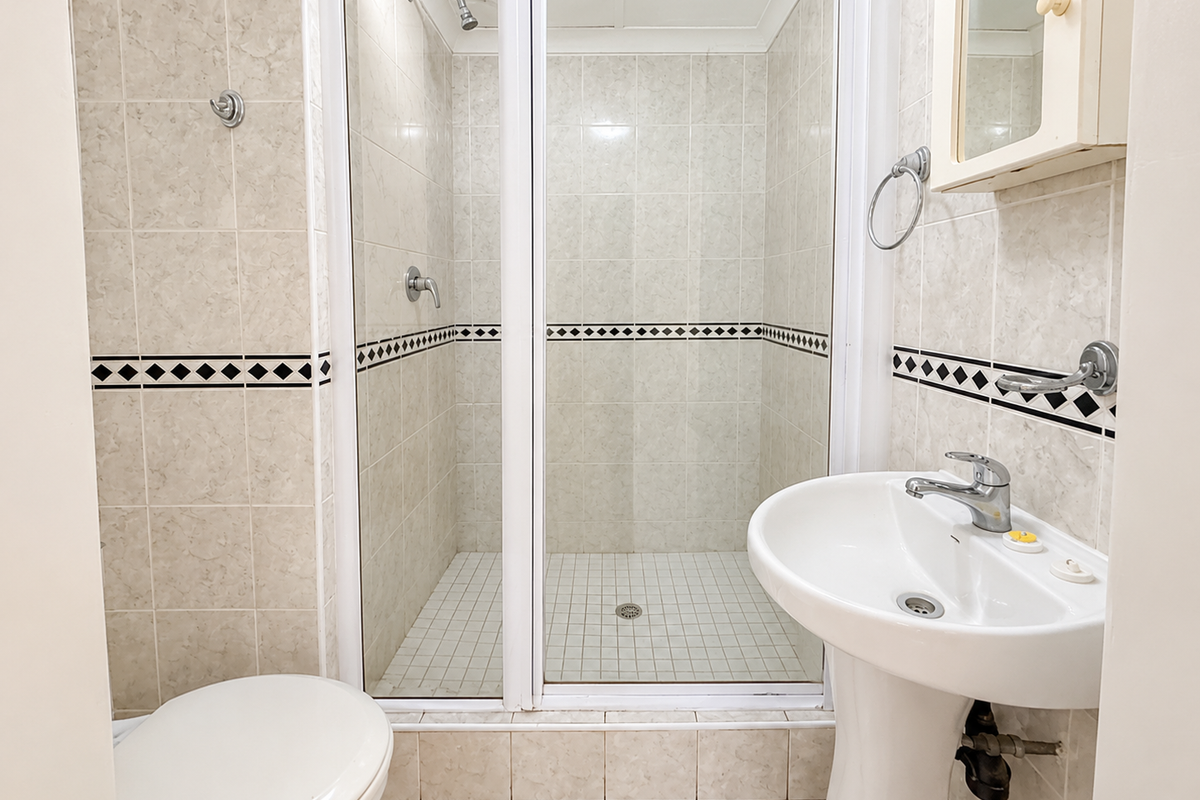 A functional bathroom featuring a pedestal washbasin, a tiled shower enclosure, and a toilet, finished with neutral wall and floor tiles.