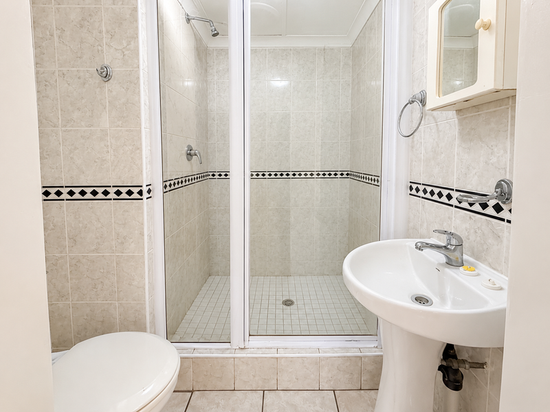 A functional bathroom featuring a pedestal washbasin, a tiled shower enclosure, and a toilet, finished with neutral wall and floor tiles.