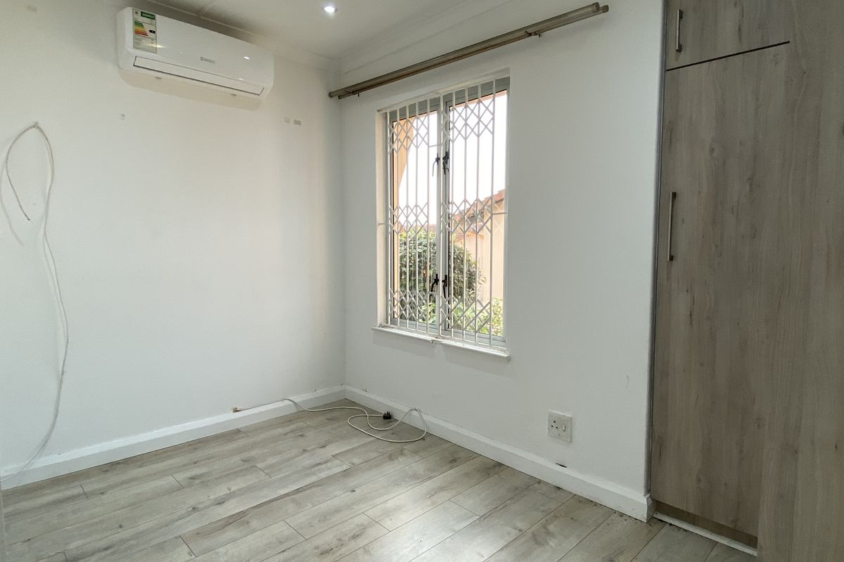 A bright, empty bedroom featuring laminate flooring, a built-in wardrobe, an air conditioning unit, and a window fitted with a security gate.