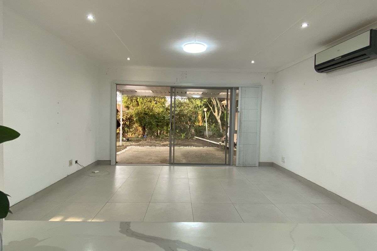 An open-plan living area featuring neutral tiled flooring, recessed lighting, and a split-system air conditioner, with direct access to the rear garden via sliding doors.