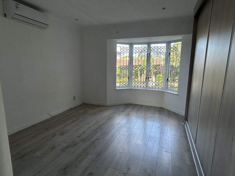 A bright, empty bedroom featuring laminate flooring, a built-in wardrobe, an air conditioning unit, and a bay window with security bars.