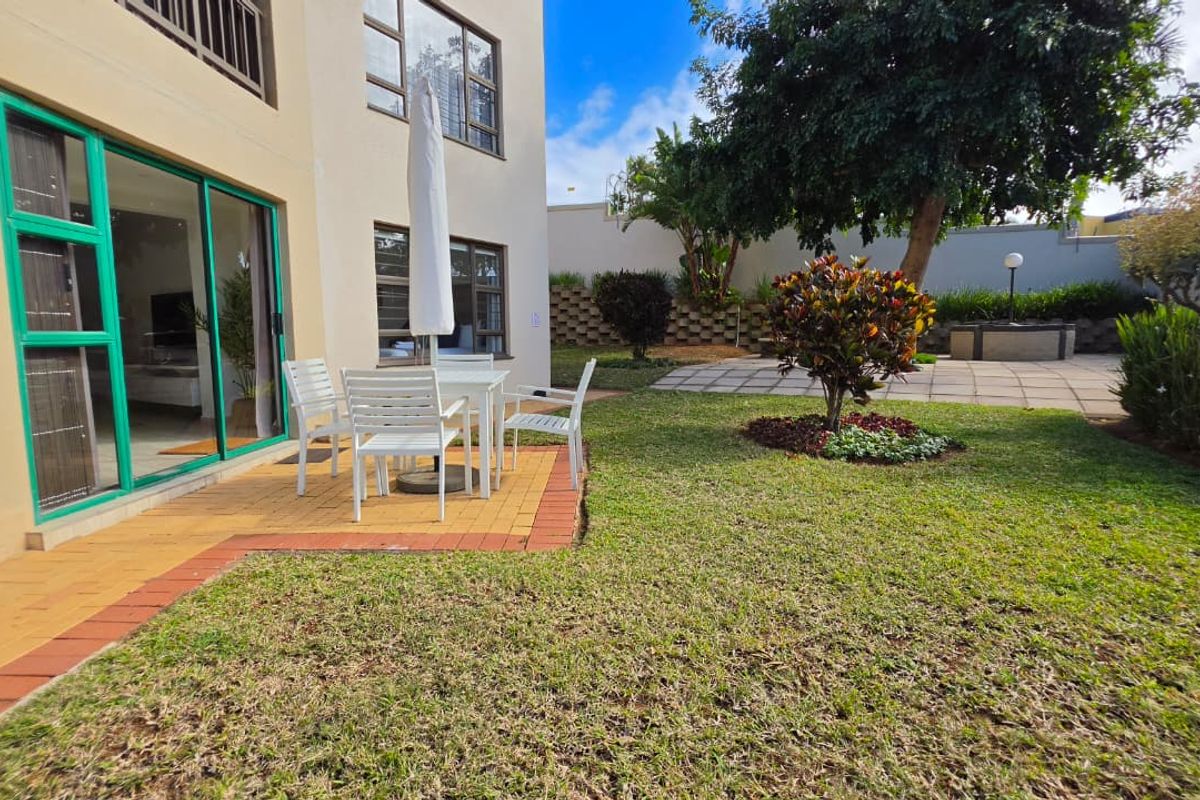 A private rear garden featuring a paved patio area, well-maintained lawn, and established landscaping, accessible via sliding glass doors from the apartment.